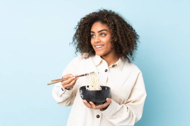 Genç Afrikalı Amerikalı bir kadın mavi arka planda izole edilmiş. Elinde çubuklarla bir kase erişte tutuyor ve yukarı bakıyor.