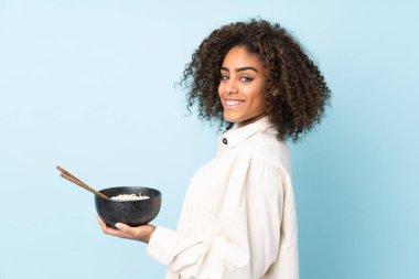 Mavi arka planda izole edilmiş genç Afrikalı Amerikalı kadın elinde çubuklarla elinde bir kase erişte tutarken gülümsüyor.