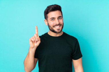 Beyaz tenli yakışıklı adam mavi arka planda izole edilmiş ve en iyinin işareti olarak parmağını kaldırıyor.