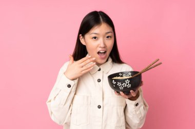Pembe arka planda izole edilmiş genç Asyalı kadın elinde yemek çubuklarıyla bir kase erişte tutuyor ve şaşırıyor.