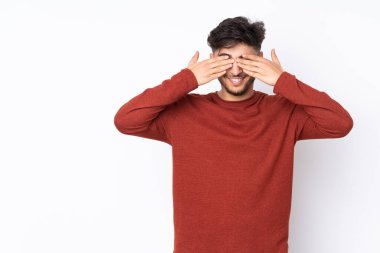 İzole edilmiş arka planda yaşayan yakışıklı Arap bir adam. Gözleri elleriyle kaplıyor ve gülümsüyor.
