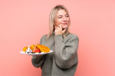 İzole edilmiş pembe arka planda waffle tutan genç sarışın kadın bir fikir üretip, bir tarafa bakıyor.