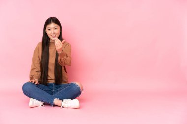 Genç Asyalı bir kadın pembe arka planda izole bir şekilde yerde oturuyor ve kendisini davet ediyor. Gelmene sevindim.