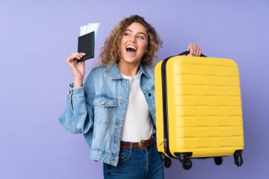 Kıvırcık saçlı genç bir kadın, mor arka planda izole edilmiş, bavul ve pasaportla tatilde.
