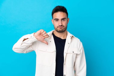 İzole edilmiş mavi arka planda genç ve yakışıklı bir adam baş parmağını aşağı gösteriyor.