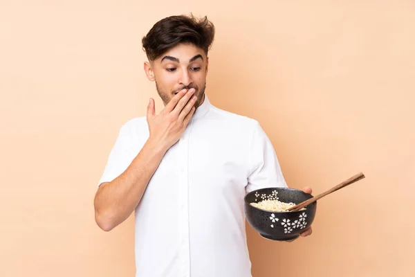 在拿着一碗用筷子做的面条时 阿拉伯帅哥面带着惊讶和震惊的表情 被米色背景隔离了 — 图库照片