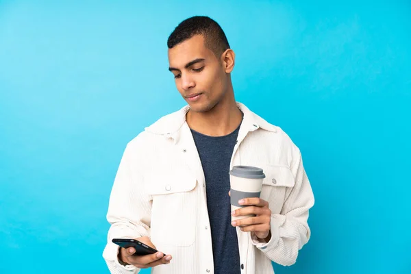 Młody Afroamerykanin Nad Odizolowanym Niebieskim Tle Trzymając Kawę Zabrania Mobilny — Zdjęcie stockowe
