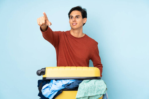 Man with a suitcase full of clothes over isolated blue background pointing away