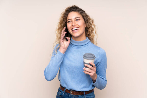 Young blonde woman with curly hair wearing a turtleneck sweater isolated on beige background holding coffee to take away and a mobile