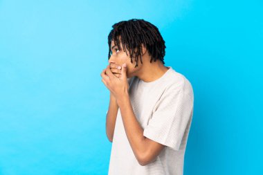 Genç Afro-Amerikalı bir adam izole edilmiş mavi arka planda ağzını kapatıyor ve yana bakıyor.