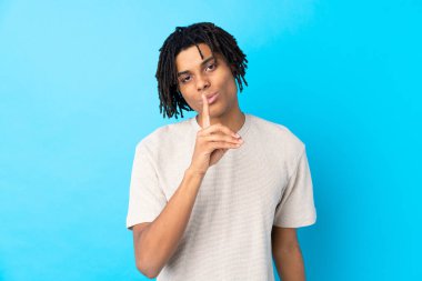 Genç Afro-Amerikalı bir adam izole edilmiş mavi arka planda sessizce hareket ettiğini gösteriyor. Parmağını ağzına sokuyor.