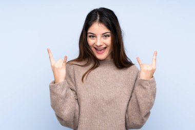 İzole edilmiş mavi arka plan üzerine kazak giyen genç esmer kadın kaya jestleri yapıyor.