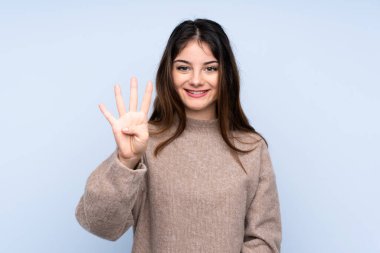 İzole edilmiş mavi arka planda süveter giyen genç esmer kadın mutlu ve parmaklarıyla dört tane sayıyor.