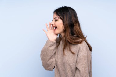 İzole edilmiş mavi arka planda süveter giyen genç esmer kadın ağzı sonuna kadar açık bağırıyor.