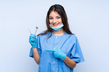 İzole edilmiş mavi arka planda aletleri tutan dişçi kadın parmak kenarı işaret ediyor.