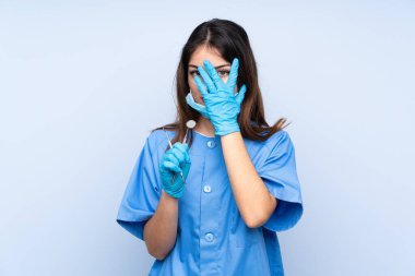 İzole edilmiş mavi arka plan üzerinde aletleri tutan kadın dişçi gözleri kapatıyor ve parmaklarından bakıyor.