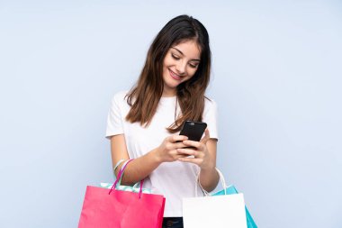 Genç esmer kadın izole edilmiş mavi arka planda elinde alışveriş torbalarıyla bir arkadaşına cep telefonuyla bir mesaj yazıyor.