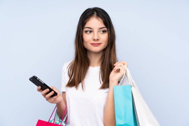 Genç esmer kadın izole edilmiş mavi arka planda elinde alışveriş çantaları ve cep telefonu tutuyor.
