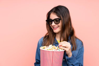 Üç boyutlu gözlüklü ve elinde koca bir kova patlamış mısırla soyutlanmış pembe arka planı olan esmer bir kadın.