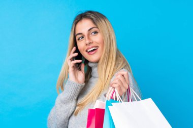 Genç Uruguaylı kadın izole edilmiş mavi arka planda elinde alışveriş çantaları ve cep telefonuyla arkadaşını arıyor.