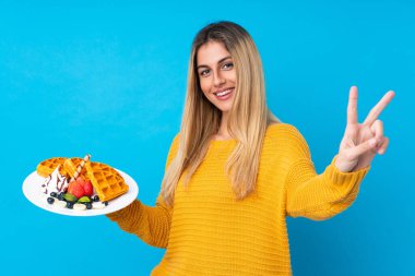 İzole edilmiş arka planda waffle tutan genç bir kadın gülümsüyor ve zafer işareti gösteriyor