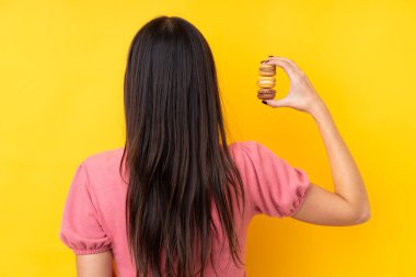 Genç esmer kadın, izole edilmiş sarı arka planda renkli Fransız kurabiyeleri tutuyor.