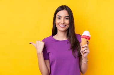 Kornet dondurmalı genç esmer kadın izole edilmiş sarı arka planda bir ürün sunmak için kenarı işaret ediyor.