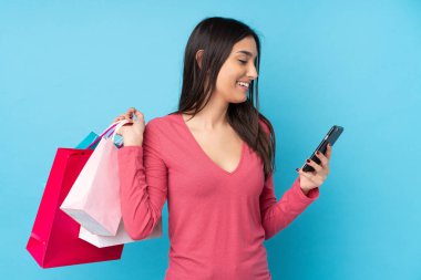 Genç esmer kadın izole edilmiş mavi arka planda elinde alışveriş torbalarıyla bir arkadaşına cep telefonuyla bir mesaj yazıyor.
