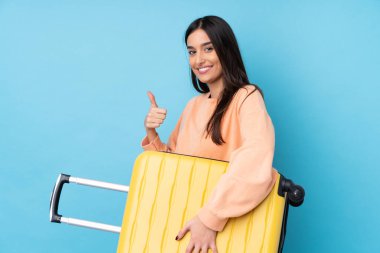 Genç esmer kadın izole edilmiş mavi arka planda seyahat valiziyle ve başparmağıyla