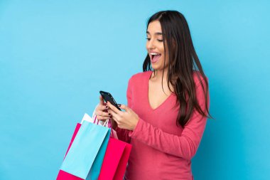Genç esmer kadın izole edilmiş mavi arka planda elinde alışveriş torbalarıyla bir arkadaşına cep telefonuyla bir mesaj yazıyor.