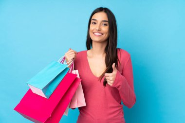 Genç esmer kadın izole edilmiş mavi arka planda elinde alışveriş çantaları ve başparmağıyla