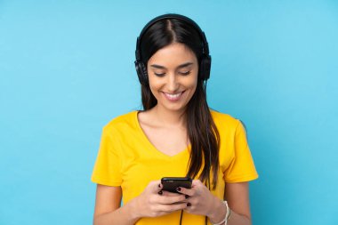 Genç esmer kadın izole edilmiş mavi arka planda müzik dinliyor ve cep telefonu arıyor.