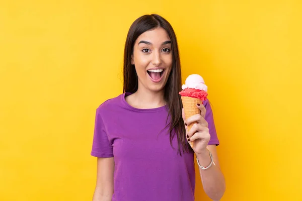 Young brunette woman with a cornet ice cream over isolated yellow background with surprise and shocked facial expression