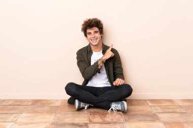 Yerde oturan genç beyaz bir adam bir ürünü sunmak için yan tarafı işaret ediyor.