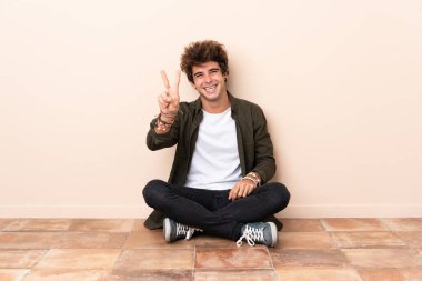 Genç, beyaz bir adam yerde oturmuş gülümsüyor ve zafer işareti gösteriyor.