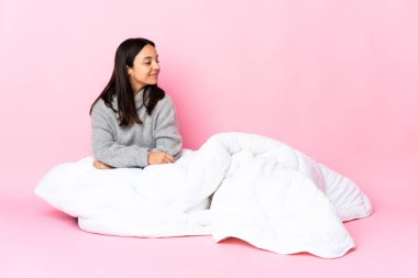 Genç, melez, pijama giymiş bir kadın yere oturmuş, yan tarafa bakıyor.