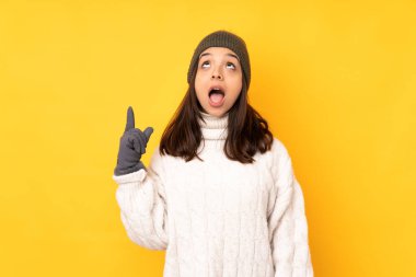 Kışlık şapkalı genç kadın izole edilmiş sarı arka planda yukarı bakıyor ve şaşırıyor.