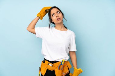 Genç elektrikçi kadın mavi arka planda izole edildi. Kafasını kaşırken şüpheleri vardı.