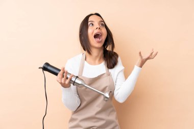 Genç esmer kız, el blendırı kullanıyor. İzole edilmiş arka planda, şaşırmış yüz ifadesi var.