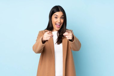 Mavi arka planda izole edilmiş genç beyaz kadın şaşırmış ve ön tarafı işaret ediyor.