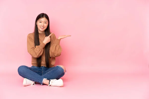年轻的亚洲女人坐在地板上 与粉红的背景隔离 手掌上握着想象的复制空间 插入一则广告 — 图库照片