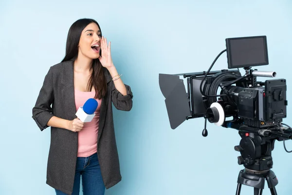 Reporter woman holding a microphone and reporting news isolated on blue background shouting with mouth wide open to the lateral