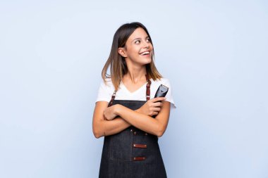 Kuaför ya da berber elbisesi olan ve elinde saç kesme makinesi olan izole edilmiş mavi arka planı olan genç bir kadın.