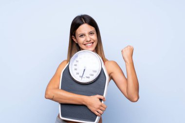 Tartı makinesi ile izole edilmiş mavi arka planda tartı makinesi olan ve zafer jesti yapan bir kız.