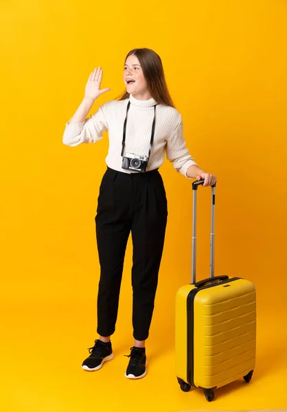 Full body of traveler teenager girl with suitcase over isolated yellow background shouting with mouth wide open