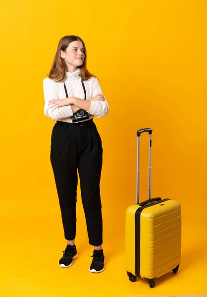 Full body of traveler teenager girl with suitcase over isolated yellow background looking to the side