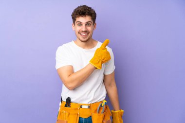 Mor arka planda izole edilmiş genç elektrikçi parmağını yana doğru gösteriyor.