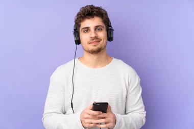 Mor arka planda izole edilmiş beyaz bir adam cep telefonuyla müzik dinliyor ve ön tarafa bakıyor.