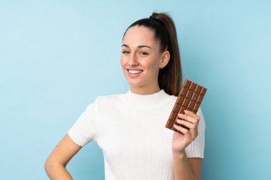 Genç esmer kadın izole edilmiş mavi arka planda çikolata tableti alıyor ve mutlu.
