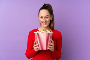 Genç esmer kadın, izole edilmiş mor arka planda elinde koca bir kova patlamış mısır tutuyor.